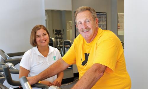 Man in yellow shirt riding bike with YMCA trainer