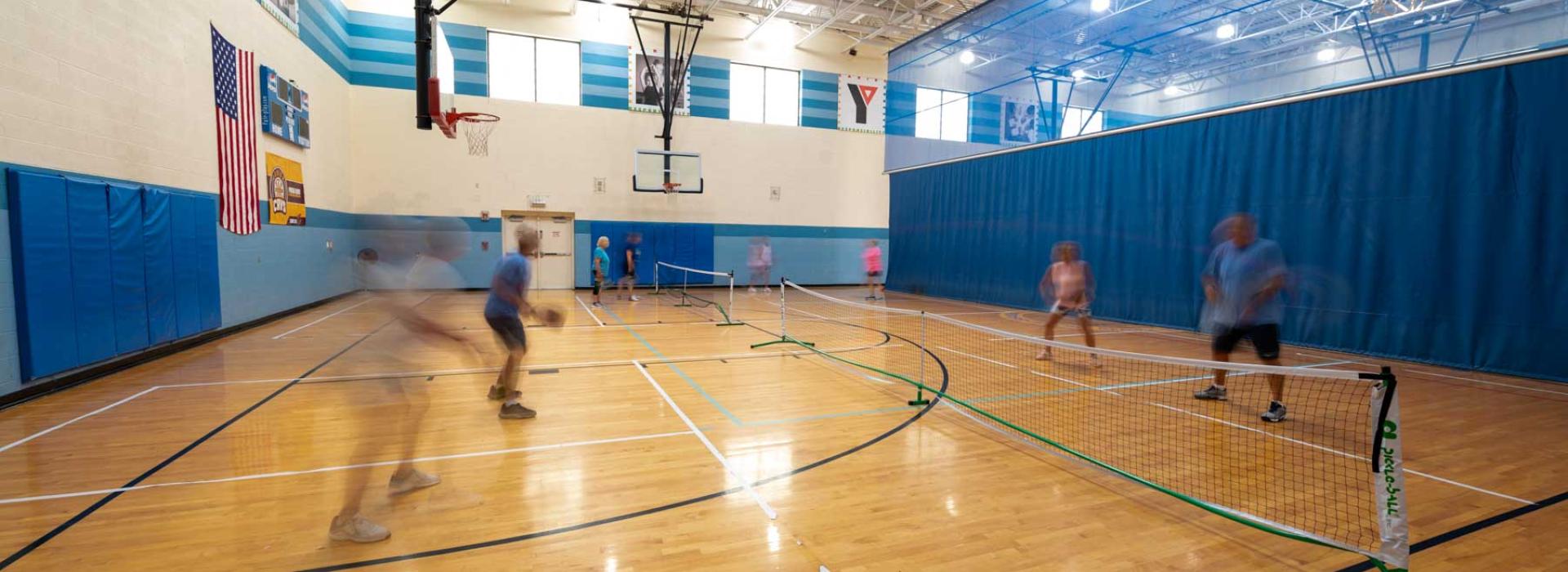 Image shows basketball court with temporary wall splitting the court. Pictured: side of gym with pickleball net set up and members playing a friendly game of pickleball. (NOT PICTURED: other side of gym is utilized for basketball at this time)